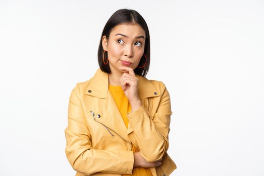 Portrait of stylish korean woman in yellow jacket, smiling thoughtful, thinking and looking up at logo or advertisement, standing over white background.