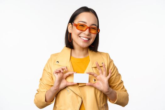 Stylish attractive asian girl in sunglasses, showing credit card and smiling, standing happy against white studio background. Copy space