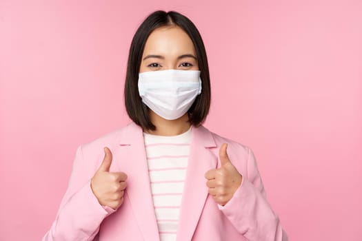 Asian businesswoman in suit and medical face mask, showing thumbs up, recommending wearing personal protective equipment in office during covid-19 pandemic, pink background.