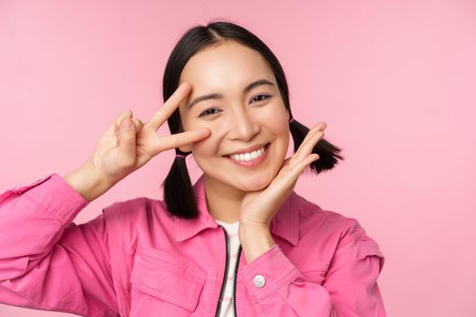 Close up of stylish asian girl smiles happy, shows peace v-sign, kawaii pose, posing against pink background. Copy space