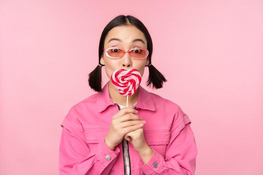 Stylish korean girl licking lolipop, eating candy and smiling, standing in sunglasses against pink background.