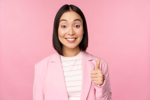 Portrait of asian businesswoman smiling satisfied, showing thumbs up, praise, like and approve, standing in suit over pink background.