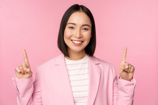 Portrait of asian businesswoman pointing fingers up and smiling, showing business company logo, information on top, standing over pink background.