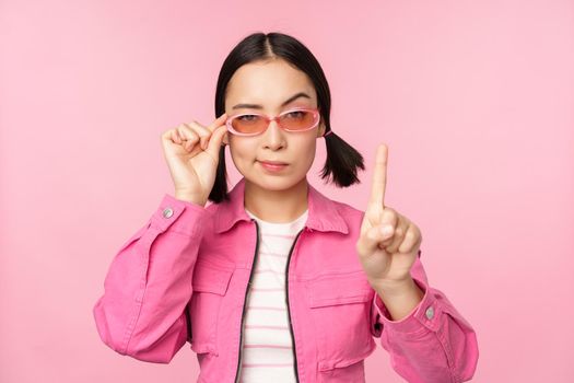 Image of asian girl with serious face, showing prohibit, scolding gesture, shaking finger pointing up, express disapproval, standing over pink background.