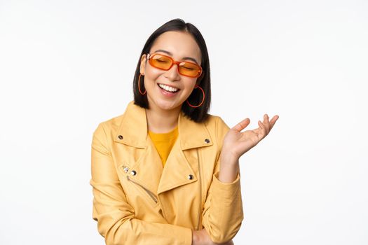 Stylish, modern korean girl in sunglasses and yellow coat, laughing and smiling, happy normal face expression, posing against white studio background.