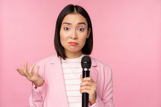 Indecisive, nervous asian business woman holding mic, shrugging and looking clueless, standing with microphone against pink background, wearing suit.