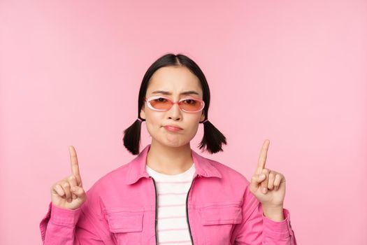 Close up of stylish korean girl, looking perplexed, pointing fingers up, showing advertisement, standing over pink background.