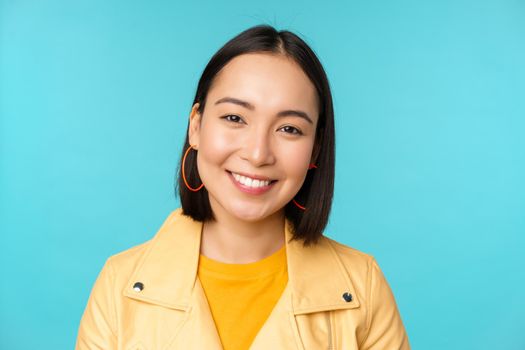 Close up portrait of smiling beautiful asian woman with white teeth, looking happy at camera, posing in yellow jacket over blue studio background.