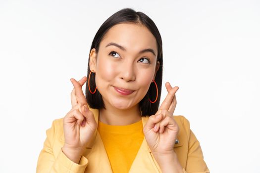 Close up portrait of hopeful asian girl wishing, cross fingers for good luck, praying and smiling, standing over white background. Copy space