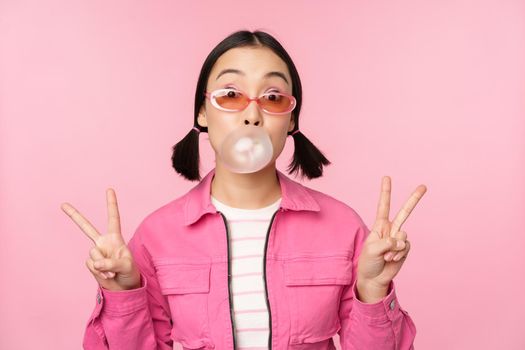 Beautiful korean girl in sunglasses, blowing bubblegum bubble and showing peace signs, standing over pink background. Copy space