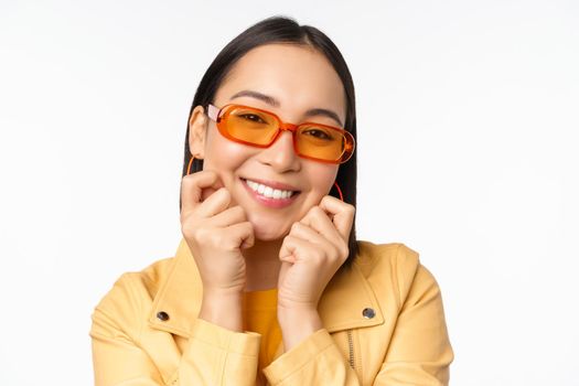 Close up portrait of trendy asian woman in sunglasses, touching her face, looking romantic, smiling at camera, standing over white background.