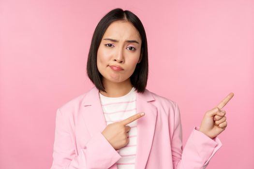 Skeptical asian businesswoman, saleswoman sulking and looking with disapproval while pointing fingers right, showing bad info, upsetting news, standing over pink background.
