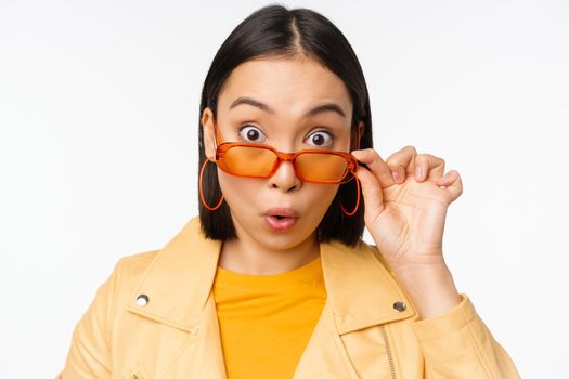 Close up portrait of asian girl looking surprised, wow face, takes off sunglasses and staring impressed at camera, standing over white background.