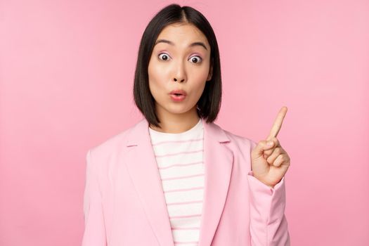 Got an idea. Young asian saleswoman, office manager raising finger, suggesting, wearing suit, posing against pink studio background.