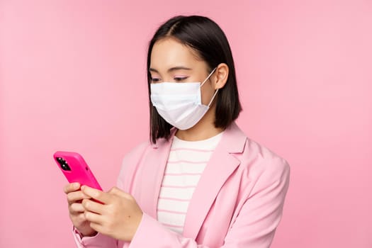 Asian businesswoman in medical face mask using mobile phone. Japenese saleswoman, corporate lady in suit, holding smartphone, standing over pink background.