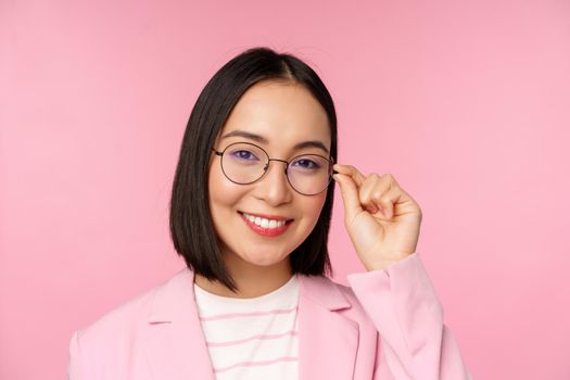 Successful asian businesswoman in glasses, smiling and looking professional at camera, wearing suit, standing over pink background.