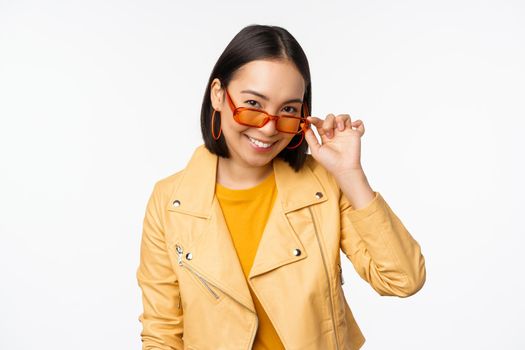 Beautiful asian girl in sunglasses, smiling and looking happy, posing in yellow jacket against white background. Copy space