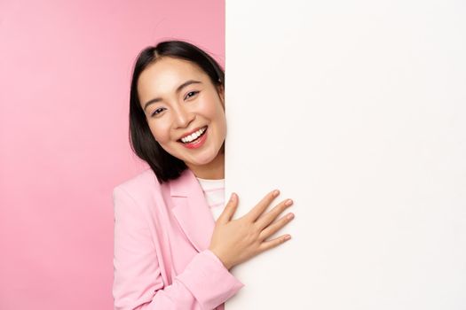 Portrait of young japanese business woman, corporate lady in suit pointing on wall with chart, showing diagram or advertisement on empty copy space, pink background.