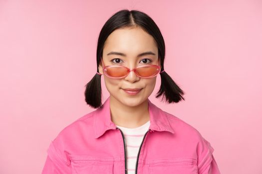 Close up of stylish korean girl in sunglasses, smiling happy, posing against pink background. People face concept.