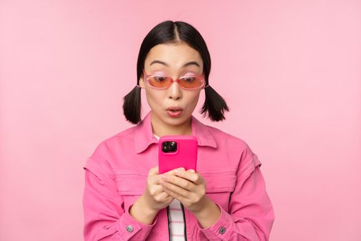 Portrait of asian girl in sunglasses using smartphone. Woman looking at mobile phone, browsing in app, standing over pink background.