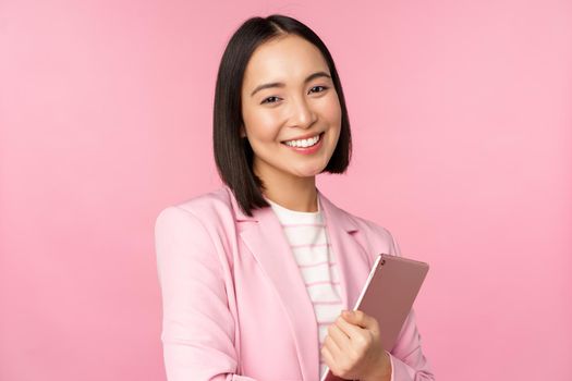 Professional smiling asian businesswoman, standing with digital tablet, wearing suit for office work, looking confident and happy, posing against pink background.