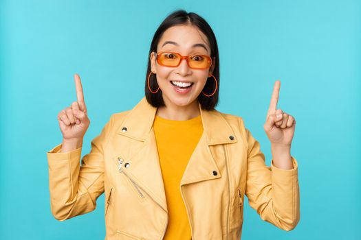 Enthusiastic asian girl in sunglasses, points fingers up, shows banner or logo on top, stands over blue background. Copy space