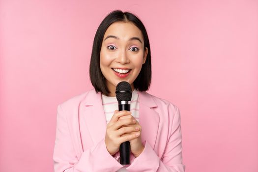 Asian businesswoman giving speech, holding microphone and smiling, standing in suit over pink background. Copy space