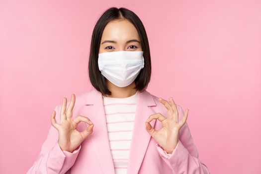 Asian businesswoman in suit and medical face mask, showing okay sign, recommending using protective equipment in office during covid-19 pandemic, pink background.