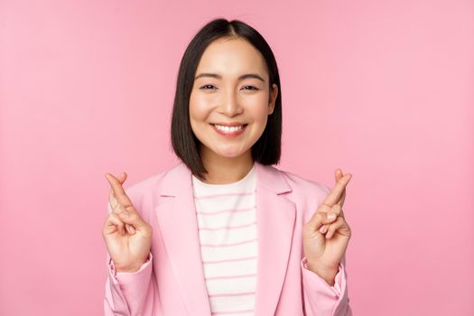 Happy asian businesswoman cross fingers for good luck, wishing, praying and hoping, smiling at camera, standing in suit over pink background.