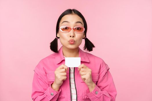 Portrait of stylish, modern asian girl, shows discount, credit card and look pleased, paying contactless, concept of shopping, standing over pink background. copy space