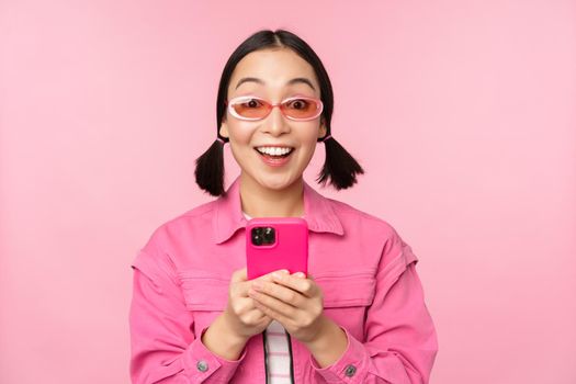 Portrait of korean girl in stylish sunglasses holding mobile phone, using smartphone app, standing over pink background. Copy space