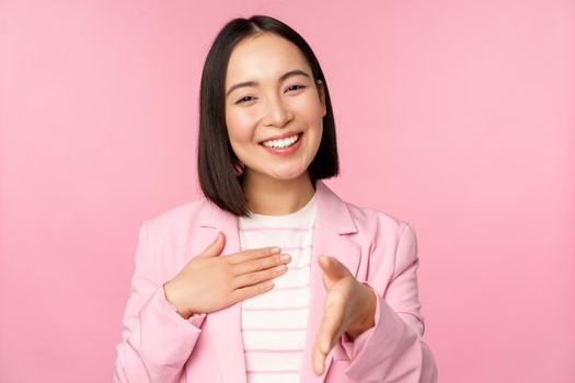 Portrait of smiling, pleasant businesswoman shaking hands with business partner, handshake, extending hand and saying hello, standing over pink background.