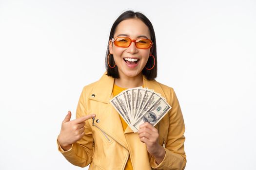 Microcredit and money concept. Stylish asian young woman in sunglasses, laughing happy, holding dollars cash, standing over white background.
