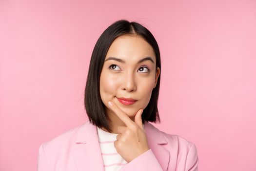 Close up portrait of young asian businesswoman thinking, smiling thoughtful and looking at upper left corner, standing over pink background.