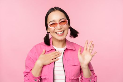 Portrait of friendly asian teen girl in sunglasses saying hello, waves her hand and smiles, greets you, hi gesture, stands over pink background.