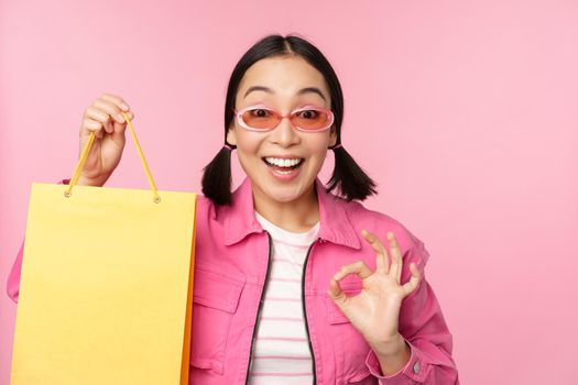 Shopping. Stylish asian girl in sunglasses, showing bag from shop and smiling, recommending sale promo in store, standing over pink background.