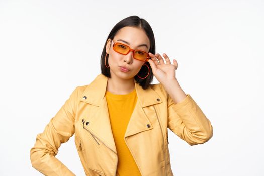 Stylish asian female model, wears sunglasses, looks confident and cool, stands over white background.