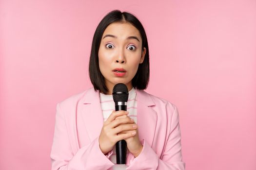 Young asian saleswoman, office lady with suit, holding microphone and looking shocked at camera, talking, giving speech, standing over pink background. Copy space