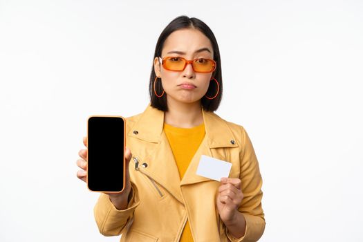 Sad asian girl in sunglasses, showing smartphone app interface, credit card, looking disappointed, standing over white background. Copy space