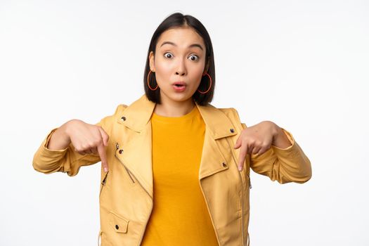 Portrait of surprised asian girl, looking amazed and impressed, pointing fingers down, showing banner with shocked face expression, white background. Copy space