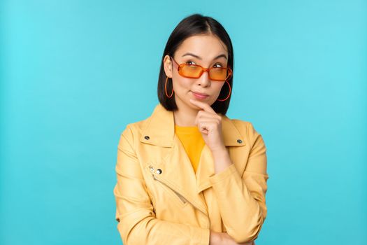 Portrait of asian woman thinking, looking thoughtful, searching ideas or solution, wearing sunglasses, standing over blue background.