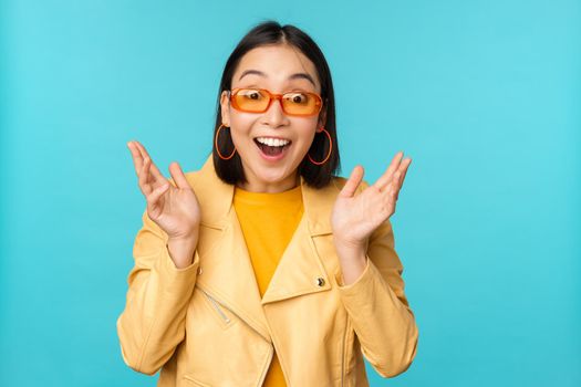 Image of enthusiastic young asian woman celebrating, triumphing, looking surprised and happy, clapping hands satisfied, standing over blue background.