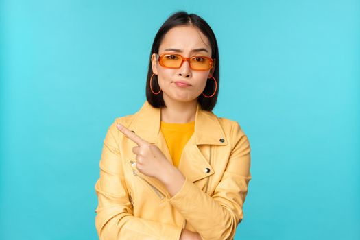 Portrait of disappointed korean woman in sunglasses, stylish girl points finger left and grimacing displeased, dislike smth, stands over blue background.