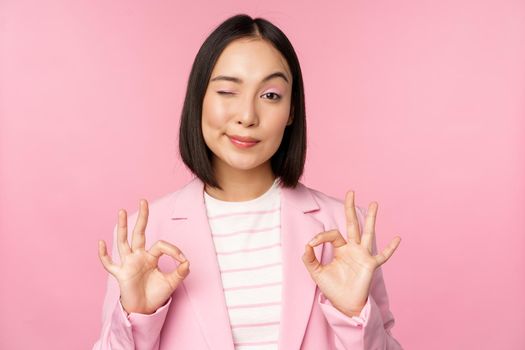 Okay, excellent. Businesswoman in corporate suit, showing ok, approval gesture, recommending smth, give positive feedback and smiling pleased, posing over pink background.
