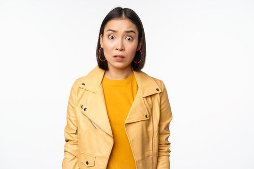 Image of shocked, confused asian woman, staring frustrated and speechless at camera, raising eyebrows and looking concerned, standing over white background.