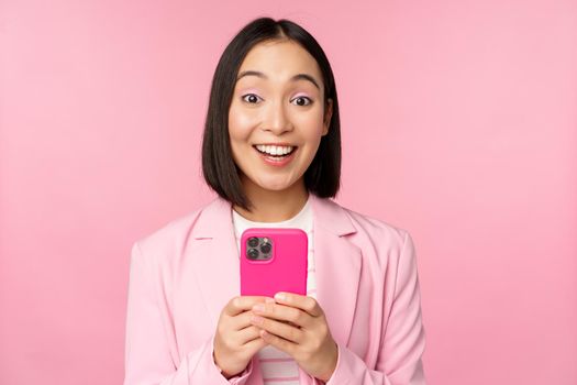 Portrait of asian girl in suit with smartphone, smiling and looking happy, standing over pink studio background. Copy space
