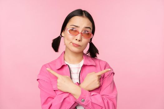 Choice. Stylish korean girl, asian female model points fingers sideways, shows two variants, product advertisement, demonstrating items, standing over pink background.