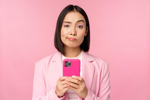 Doubtful businesswoman in suit, holding smartphone, grimacing, making skeptical face expression, standing over pink background.