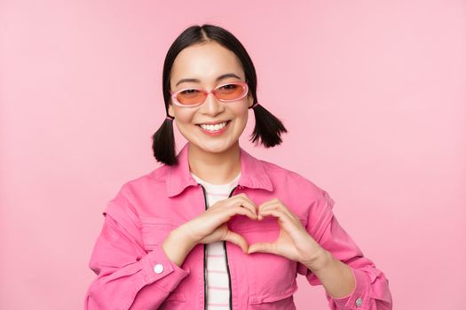 Lovely korean female model in trendy sunglasses, shows heart, care sign, I love you gesture, stands over pink background.