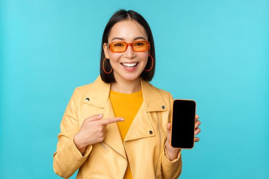 Image of young stylish asian female model in trendy sunglasses, showing mobile phone screen, application on smartphone interface, standing over blue background.